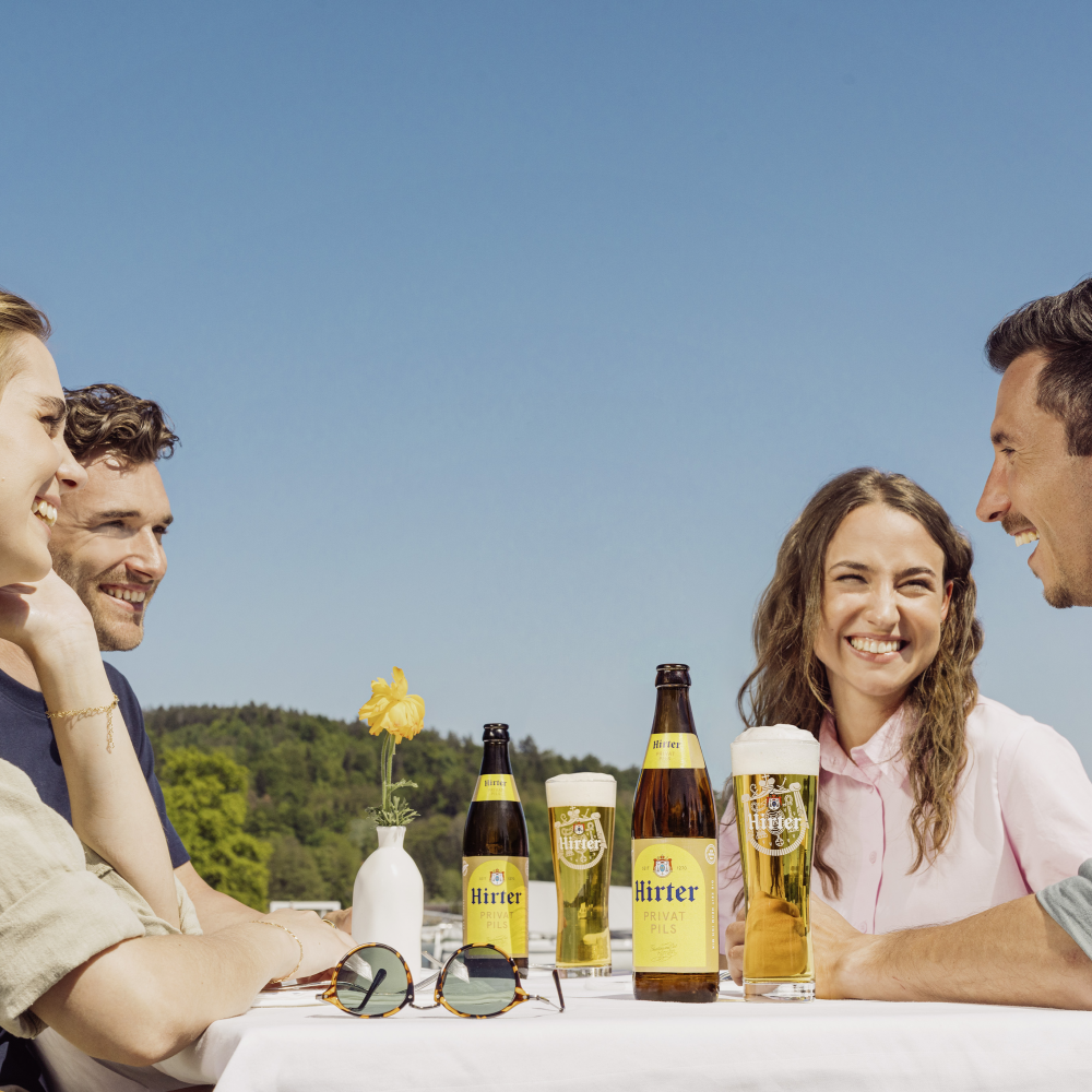 4 Menschen sitzen plaudernd auf einem Tisch zusammen und mittig stehen Hirter Bierflaschen und Gläser | © Privatbrauerei Hirt